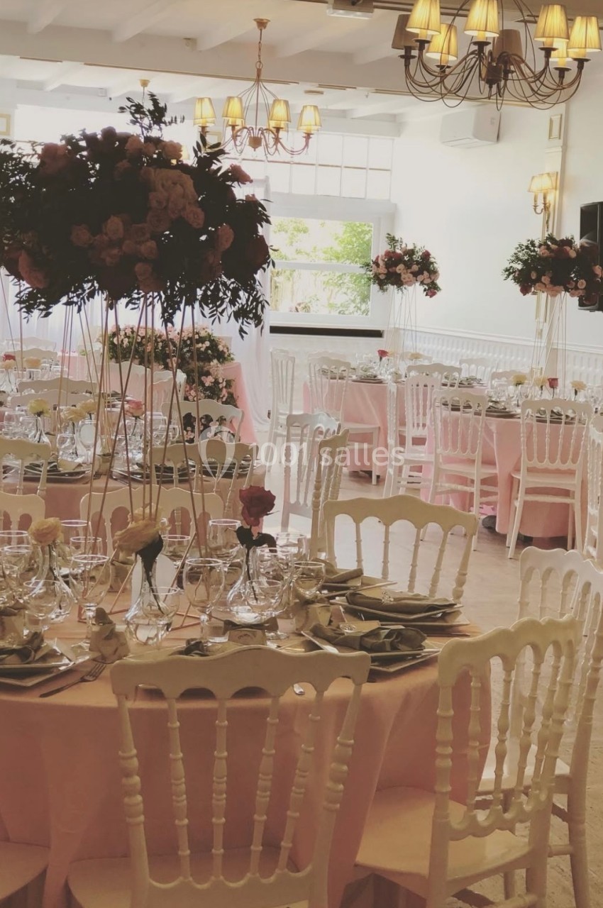 Salle de réception décorée avec des tables rondes, nappes roses, chaises blanches et centres de table fleuris.