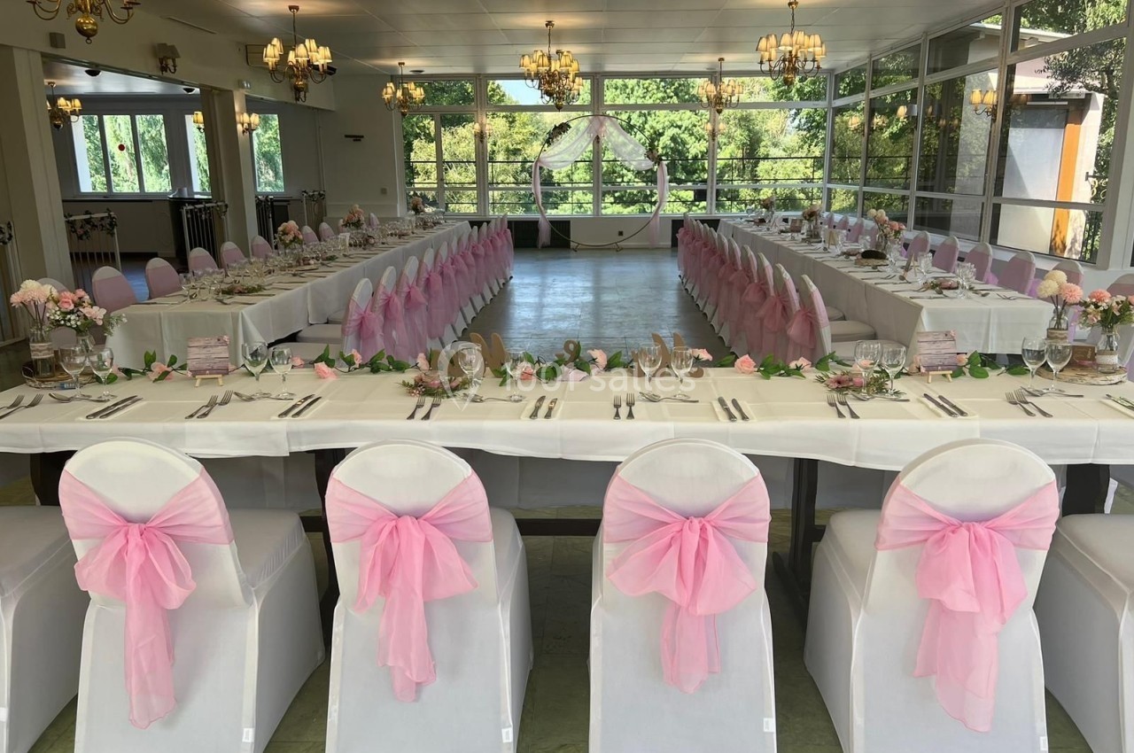 Salle de réception décorée pour un mariage, avec des tables en U, nappes blanches et chaises ornées de nœuds roses.