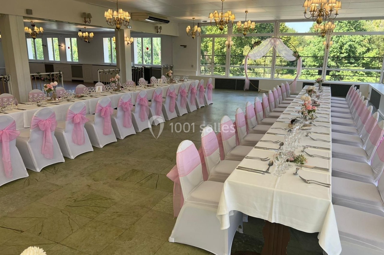 Salle de réception décorée pour un mariage, avec chaises blanches ornées de nœuds roses et tables dressées.