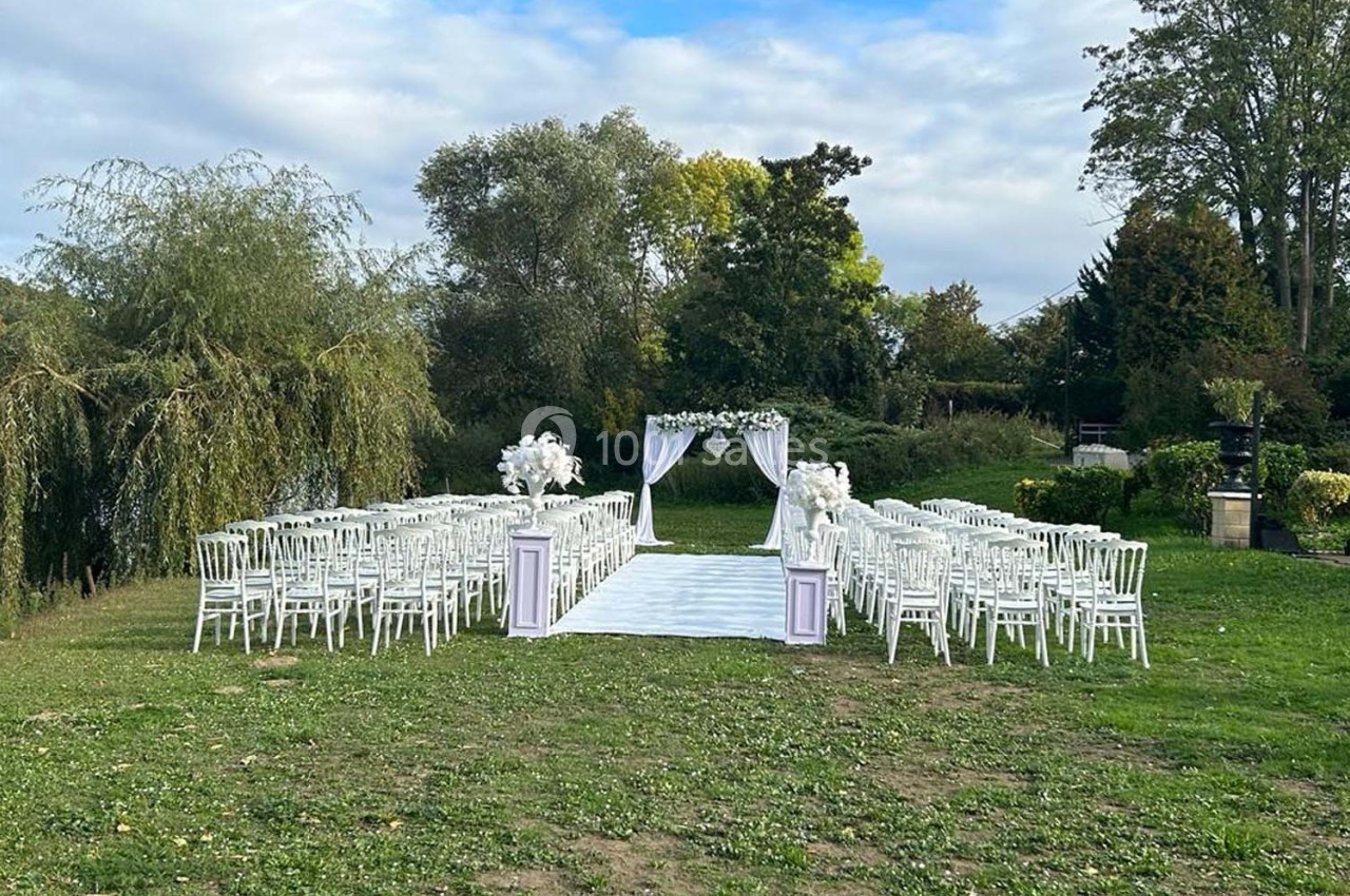 Allée centrale bordée de chaises blanches menant à une arche décorée pour une cérémonie en plein air dans un jardin…