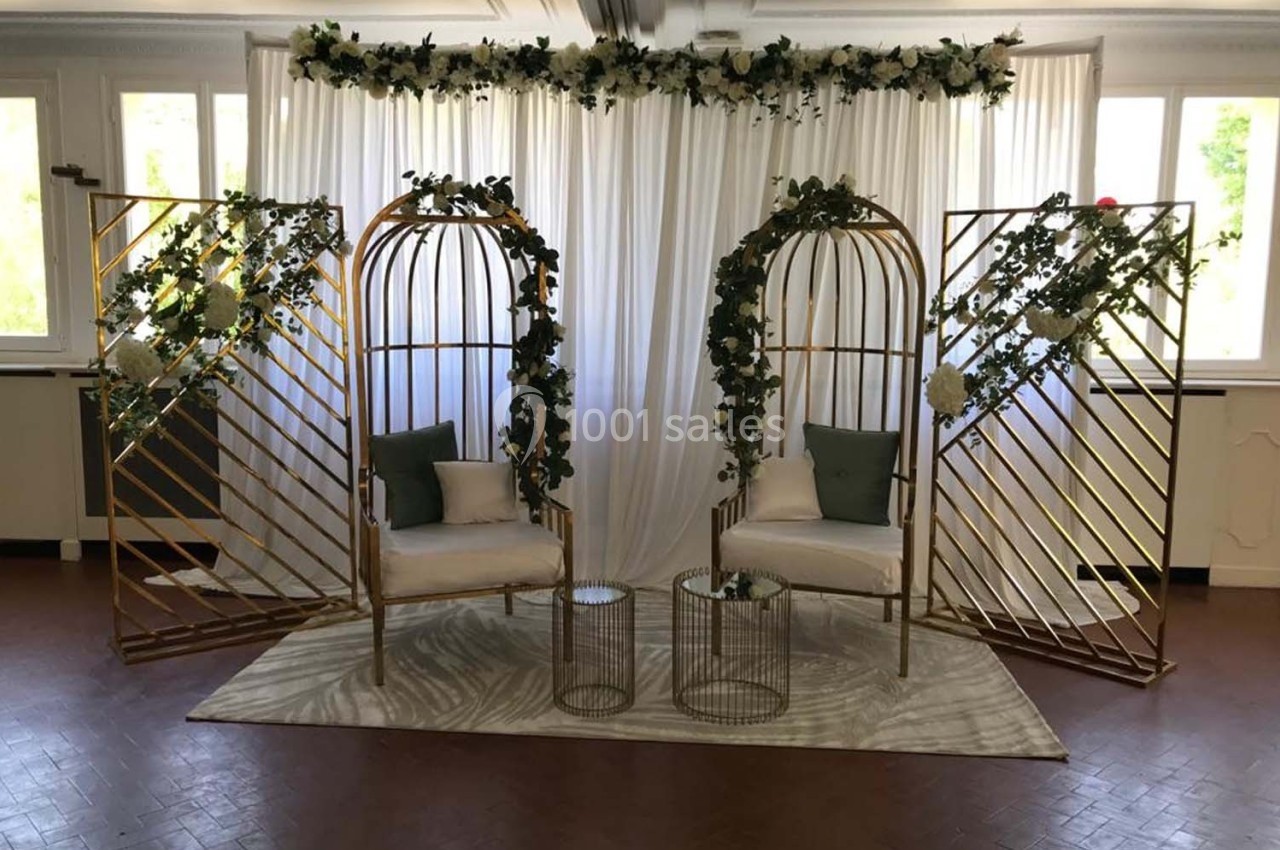 Deux fauteuils blancs entourés de structures dorées décorées de feuillage, devant un rideau blanc.