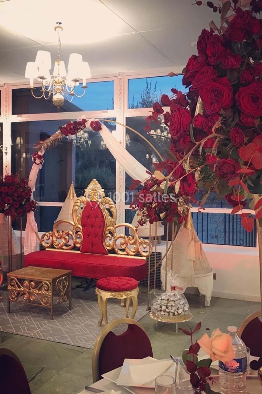 Salle décorée pour un événement avec un trône rouge et or, des arches fleuries de roses rouges et des tables dressées.