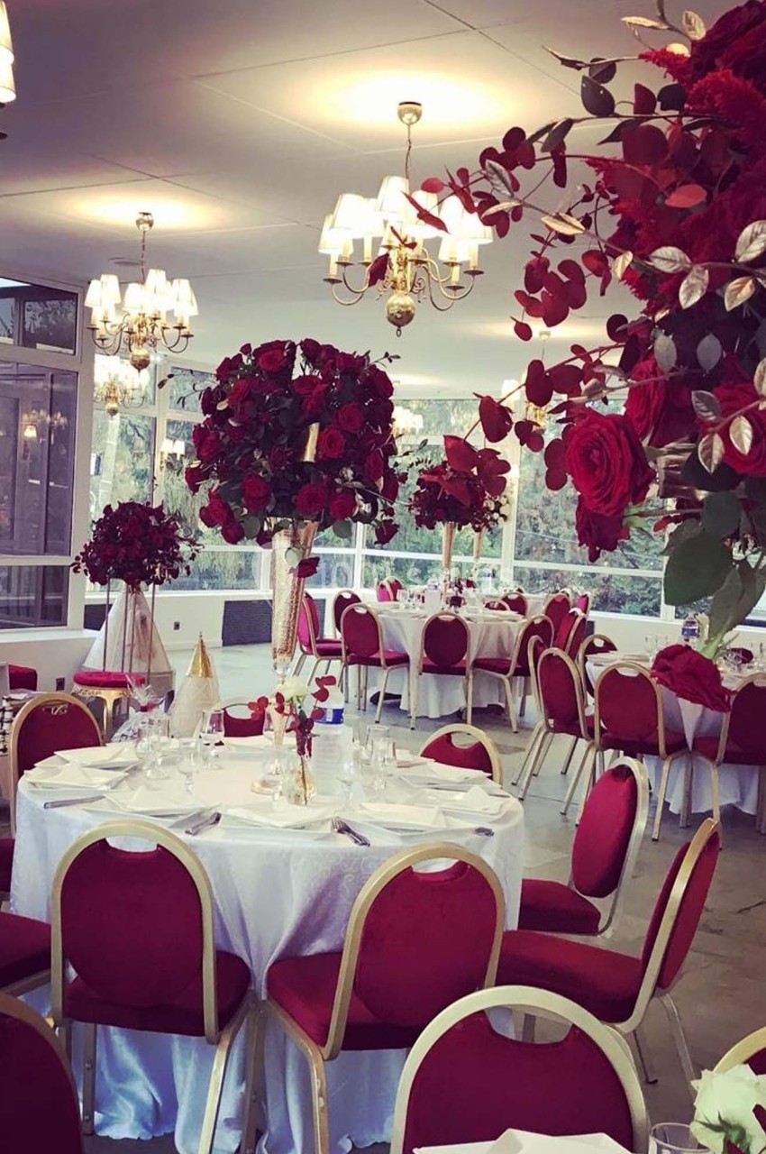 Salle de réception décorée avec des tables rondes, nappes blanches, chaises rouges et arrangements floraux rouges.
