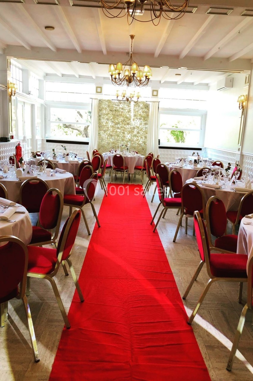 Salle de réception lumineuse avec tables dressées et chaises rouges, traversée par un tapis rouge central.