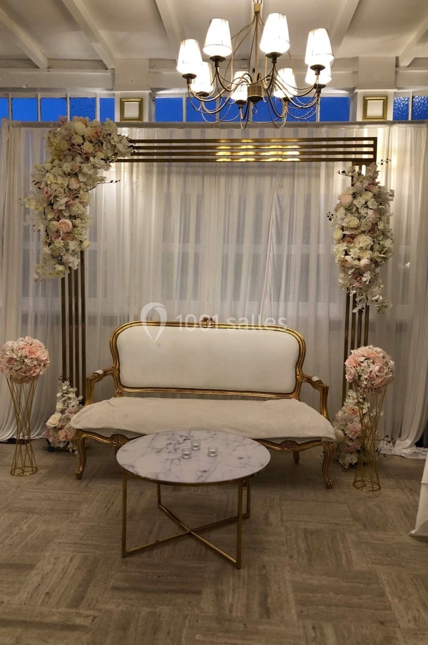 Canapé blanc et doré devant un rideau blanc, entouré d'arches fleuries, sous un lustre dans une salle lumineuse.