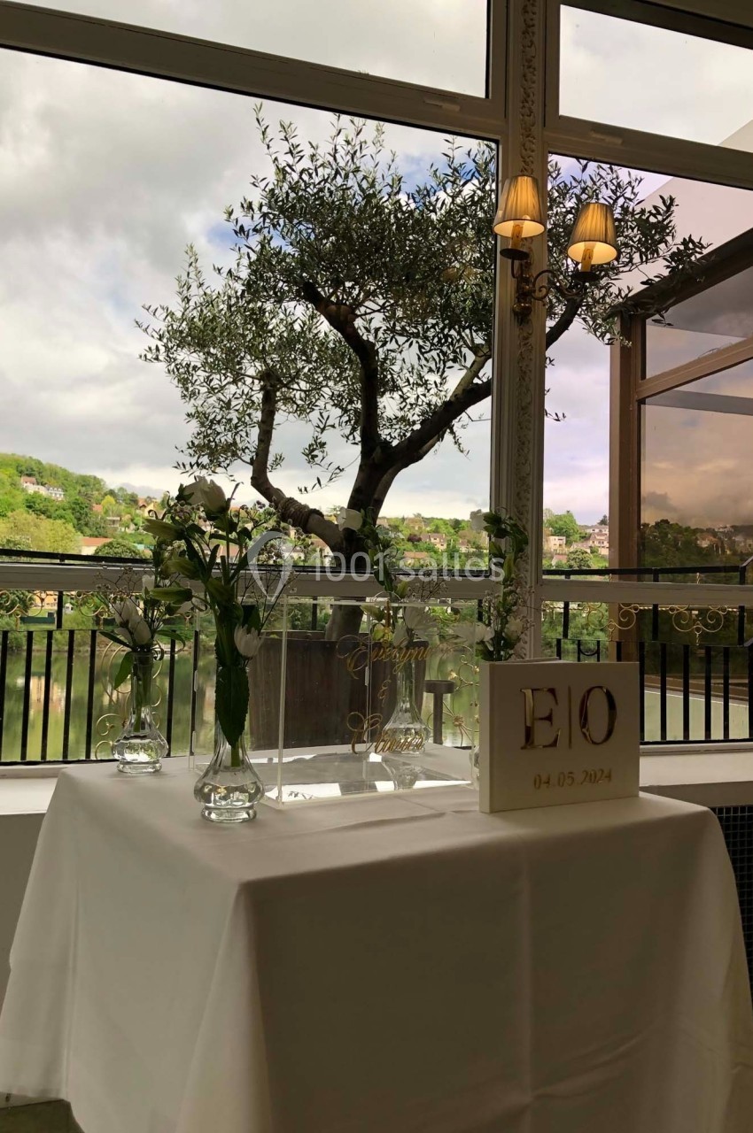 Table décorée avec des fleurs blanches et un panneau personnalisé, devant une baie vitrée avec vue sur un arbre et un…
