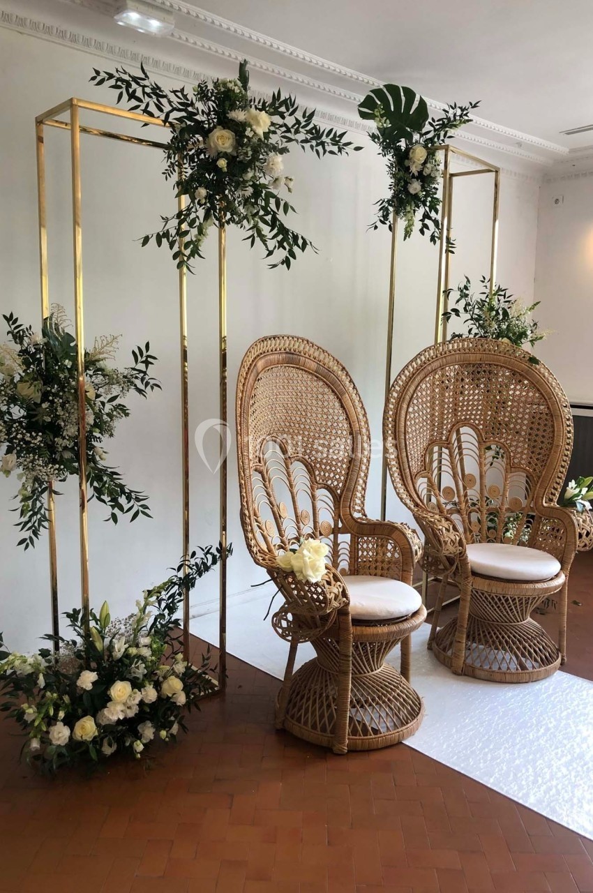 Deux fauteuils en rotin entourés de structures dorées décorées de fleurs blanches et de feuillage dans une salle lumineuse.
