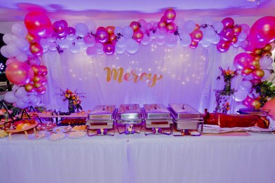 Buffet décoré avec des ballons roses et blancs, plats variés, et un cochon rôti sur une table drapée de blanc.