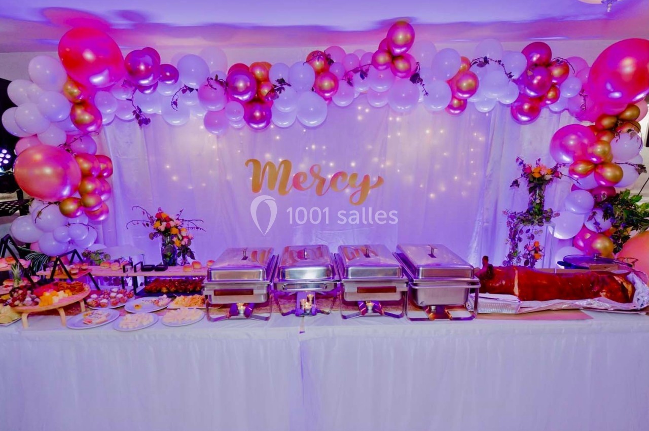 Buffet décoré avec des ballons roses et blancs, plats variés, et un cochon rôti sur une table drapée de blanc.