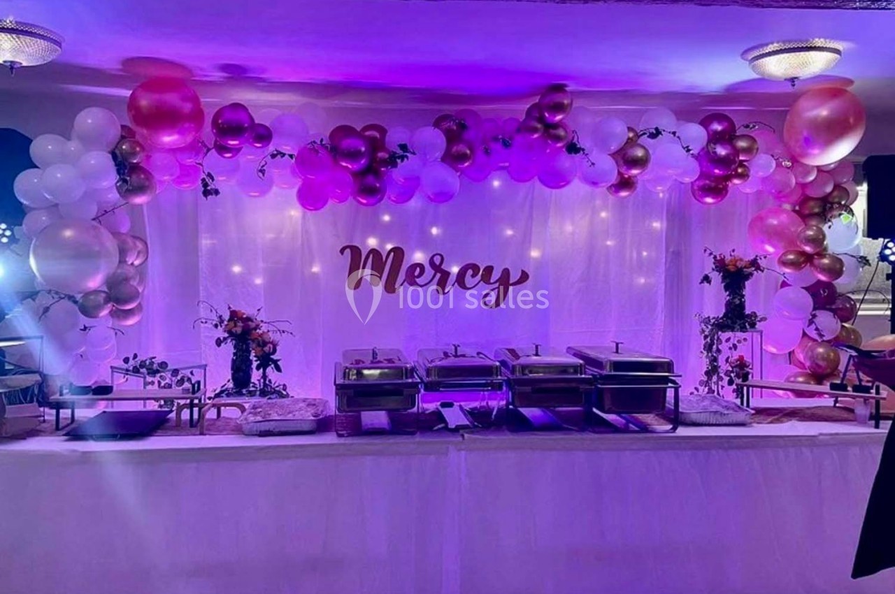 Décoration de buffet avec ballons roses et blancs, guirlandes lumineuses et le mot ’Mercy’ en arrière-plan.