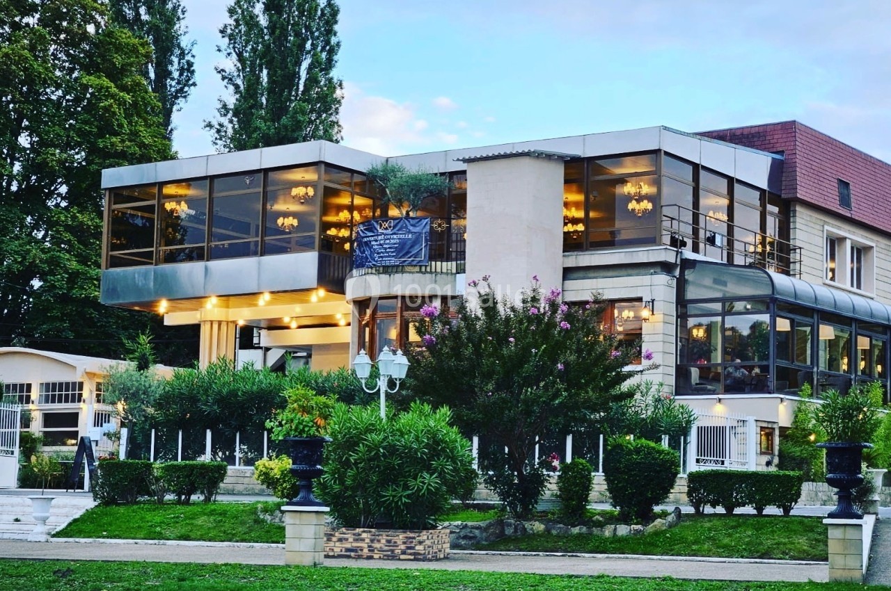 Bâtiment moderne avec de grandes baies vitrées entouré de verdure et éclairé par des luminaires en soirée.