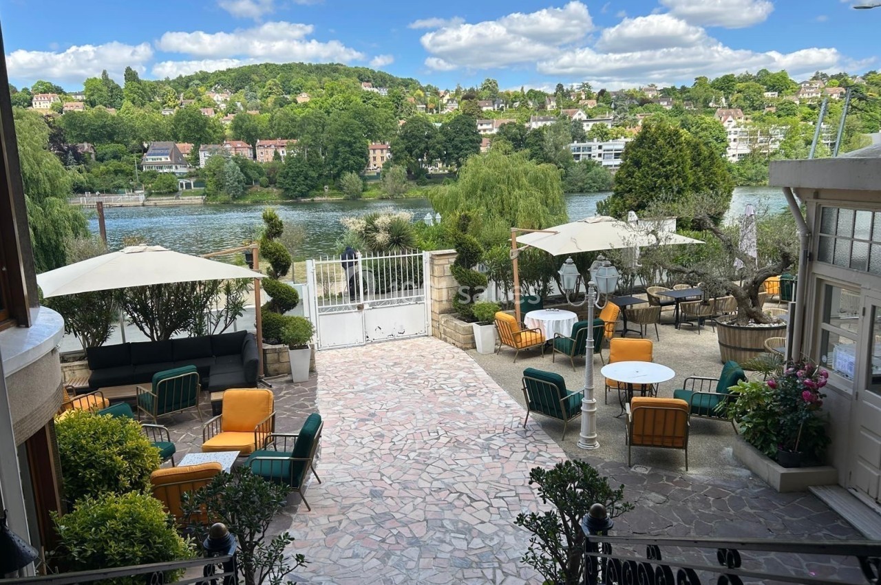 Terrasse aménagée avec tables, chaises et parasols, offrant une vue sur une rivière et des collines verdoyantes.