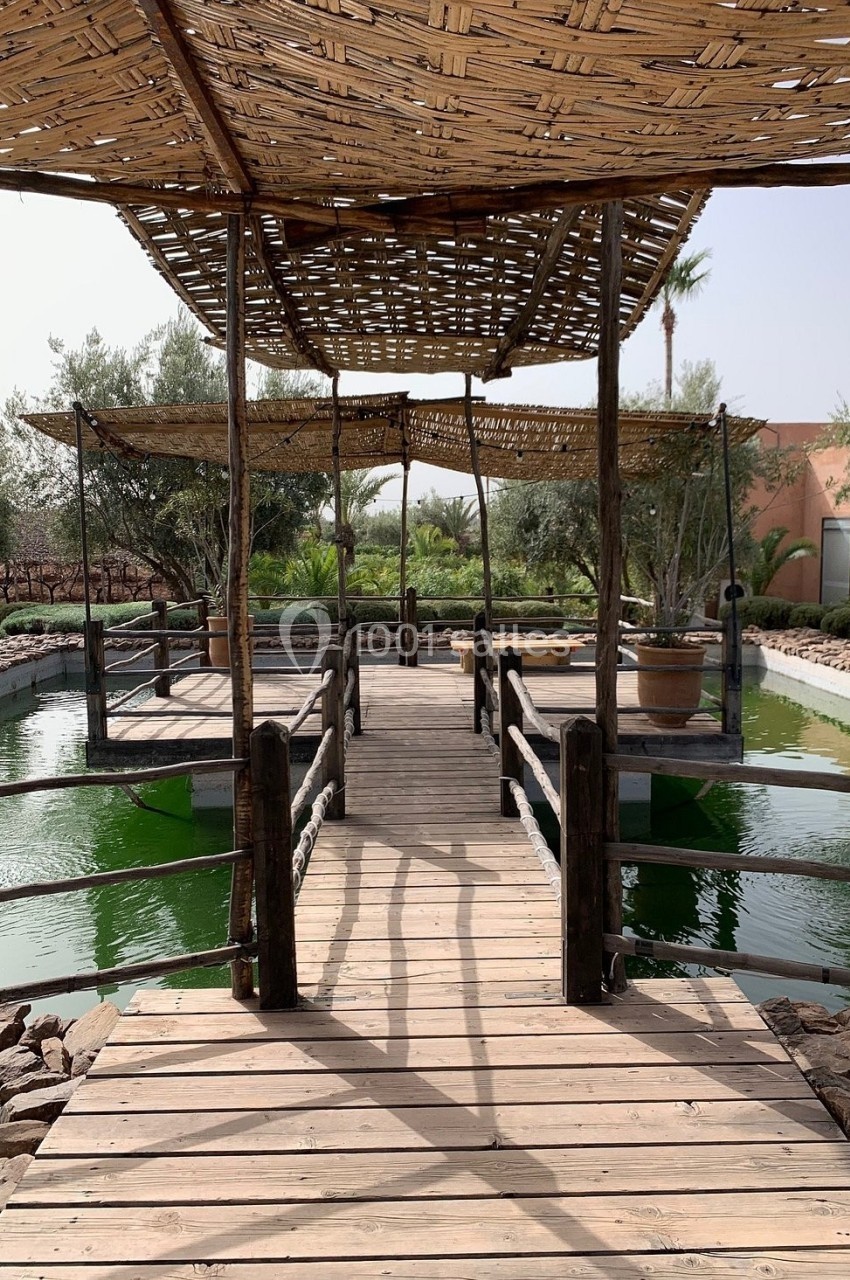 Passerelle en bois avec pergola en roseaux traversant un bassin entouré de végétation et d'oliviers.