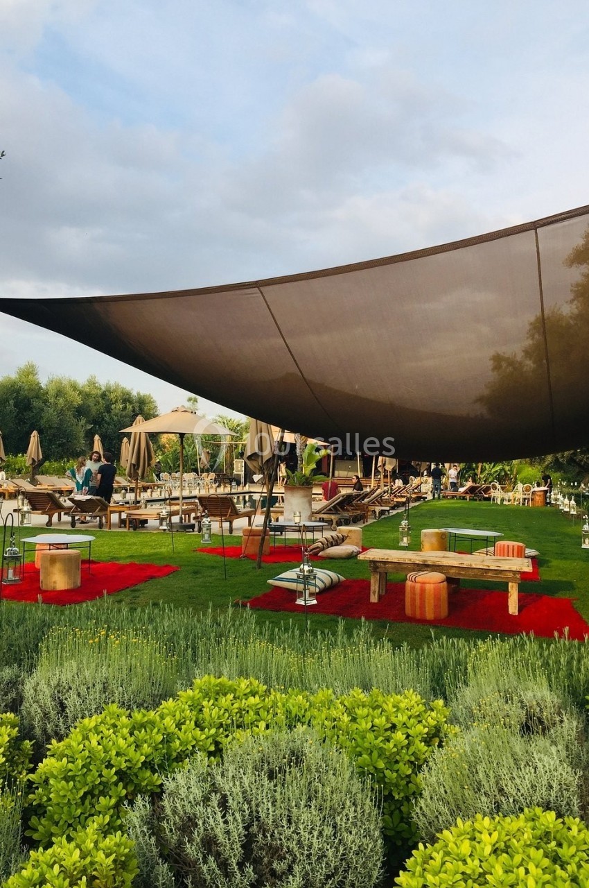 Espace extérieur avec pelouse, tapis rouges, mobilier bas, voiles d'ombrage et chaises longues près d'arbres et buissons.