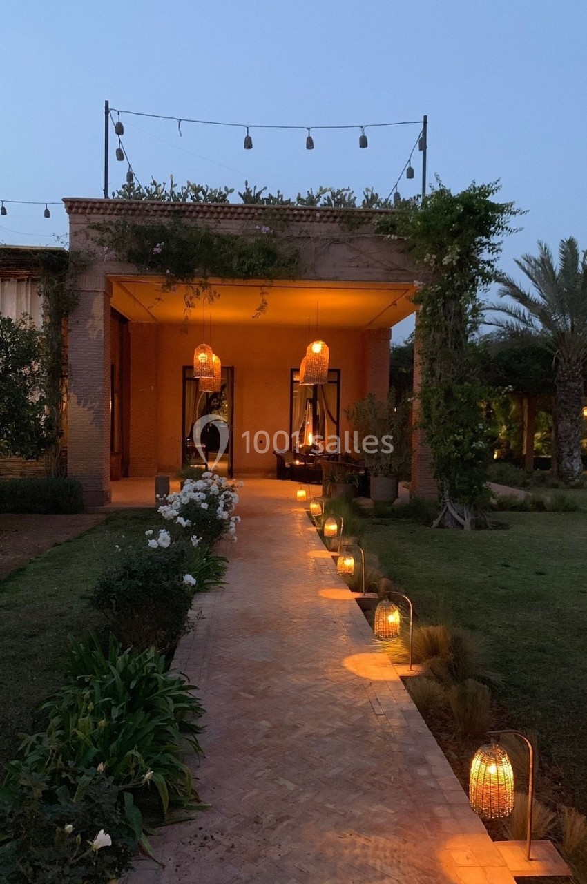 Allée éclairée par des lanternes menant à une terrasse illuminée au crépuscule, entourée de végétation.