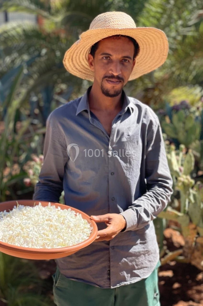 Un homme portant un chapeau de paille tient un plateau rempli de petites fleurs blanches dans un jardin ensoleillé.