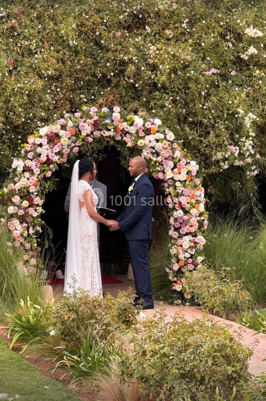 Un couple échange ses vœux de mariage devant une arche fleurie dans un jardin verdoyant.