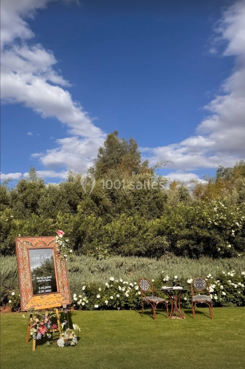 Panneau décoratif entouré de fleurs, avec deux chaises et une table sur une pelouse, sous un ciel bleu parsemé de nuages.