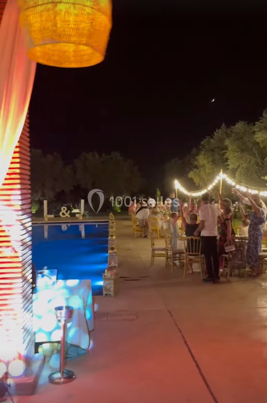 Des invités rassemblés autour de tables éclairées par des guirlandes lumineuses près d'une piscine, en soirée.