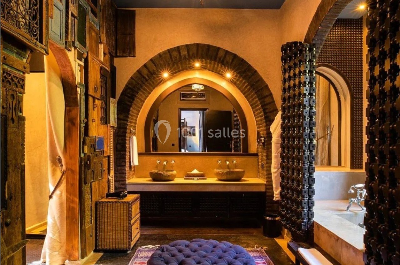 Salle de bain au style marocain avec arches, tapis coloré, pouf capitonné et éclairage suspendu.