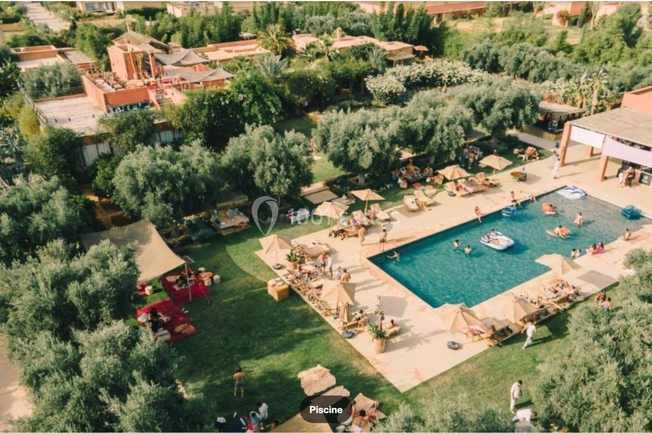 Vue aérienne d'une piscine entourée de transats, parasols, arbres et personnes profitant d'un cadre verdoyant.