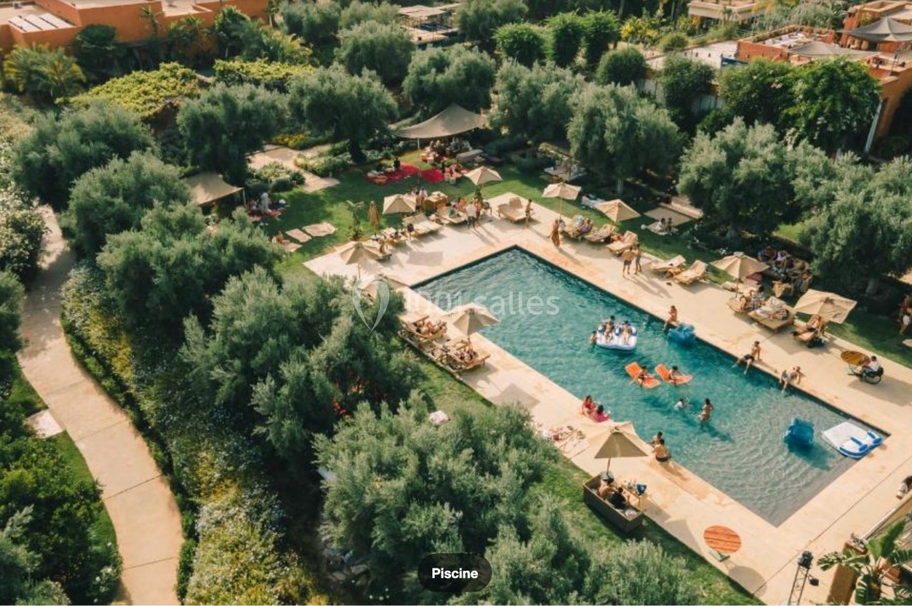 Vue aérienne d'une piscine entourée de chaises longues, parasols et végétation luxuriante, avec des baigneurs.