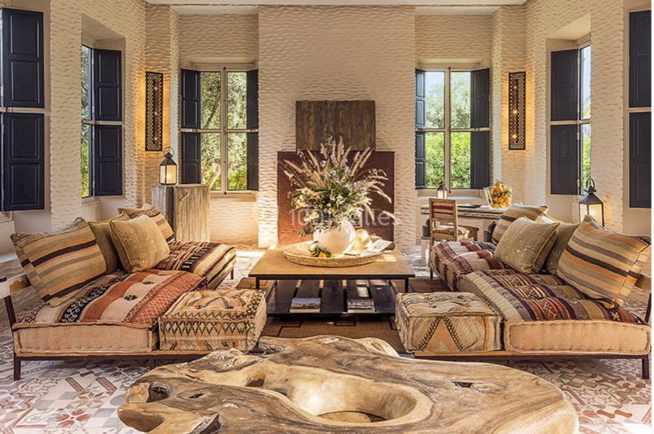 Salon lumineux avec canapés en tissu à motifs, table basse en bois sculpté et grandes fenêtres noires.