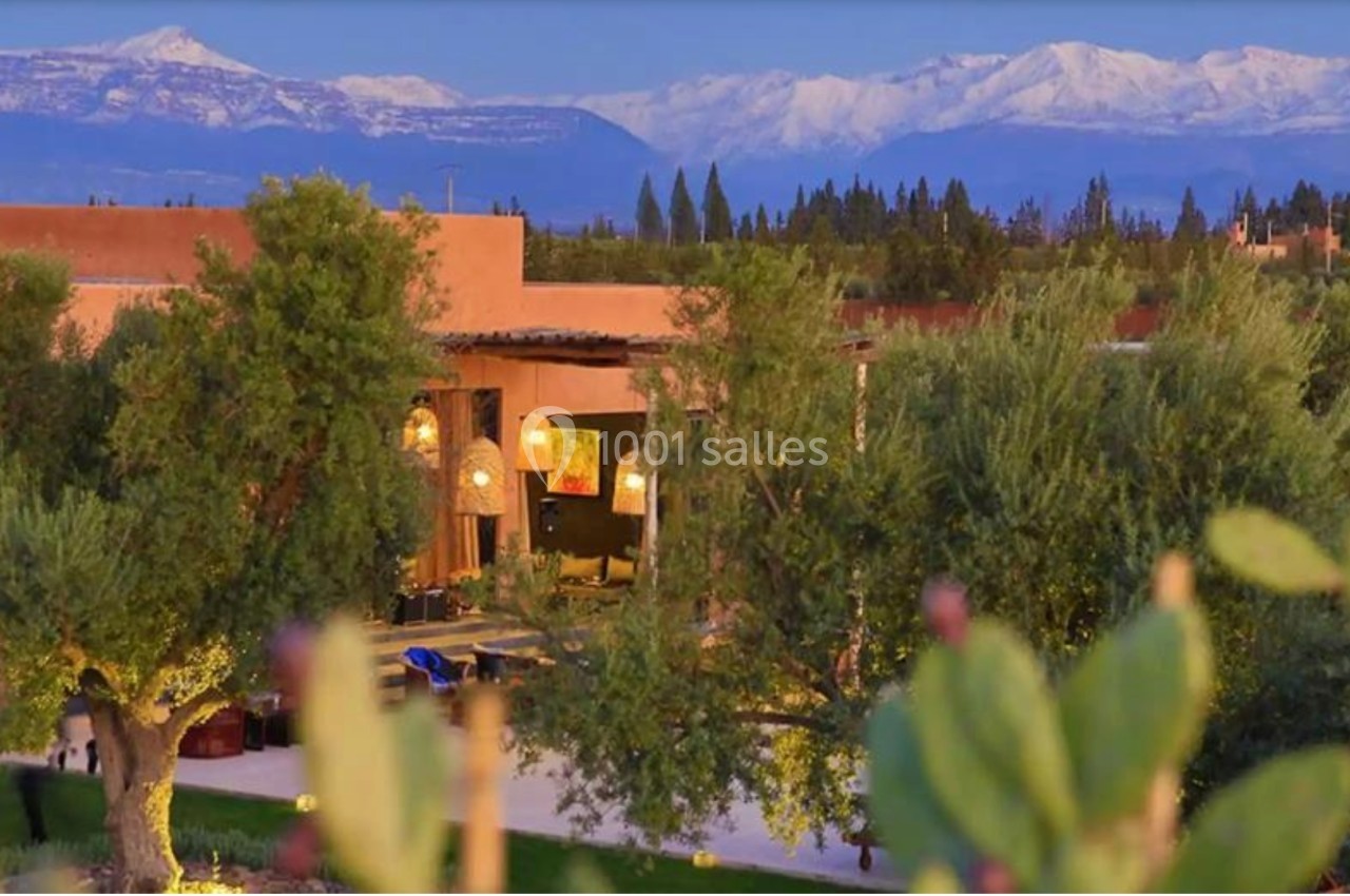 Vue d'une maison en terre cuite entourée d'arbres, avec des montagnes enneigées en arrière-plan sous un ciel clair.