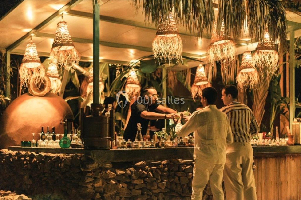 Un barman sert deux clients dans un bar extérieur décoré de lampes suspendues et de végétation tropicale.