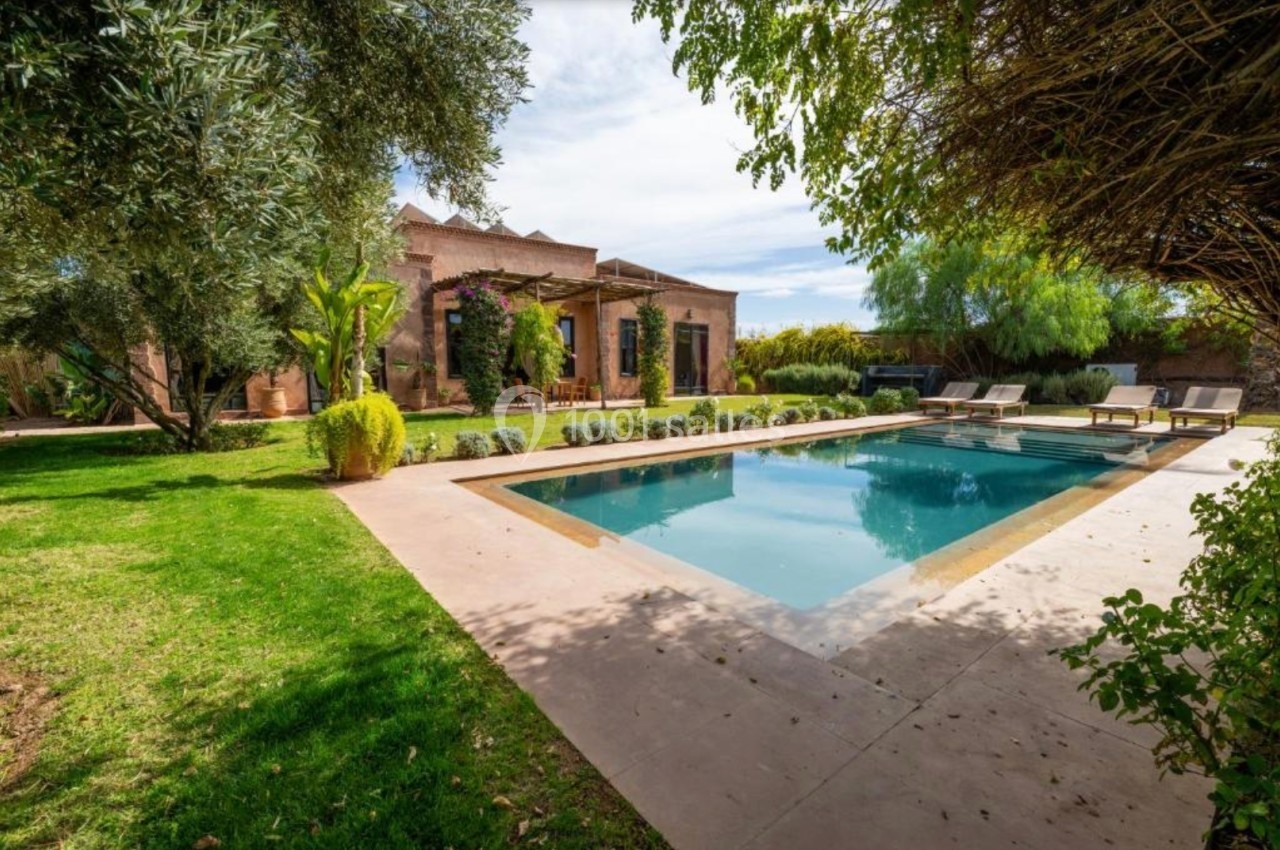 Piscine extérieure entourée de verdure et d'arbres, avec une maison en arrière-plan sous un ciel dégagé.