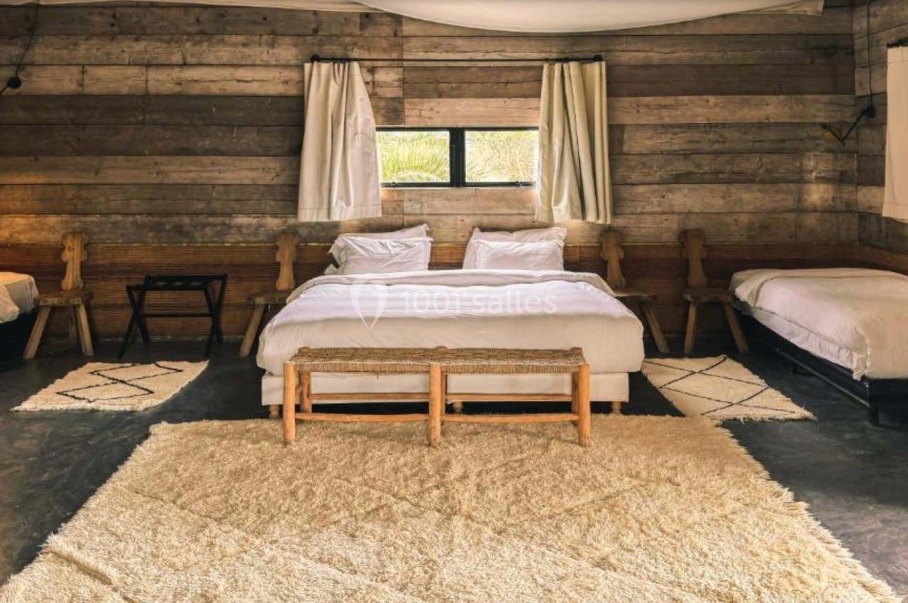 Chambre rustique avec murs en bois, lit double central, deux lits simples, tapis beige et lumière naturelle.