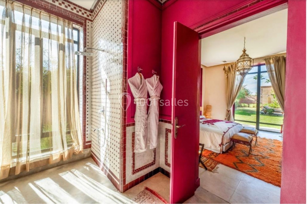 Salle de bain avec douche ouverte, peignoirs accrochés, et vue sur une chambre lumineuse donnant sur un jardin.