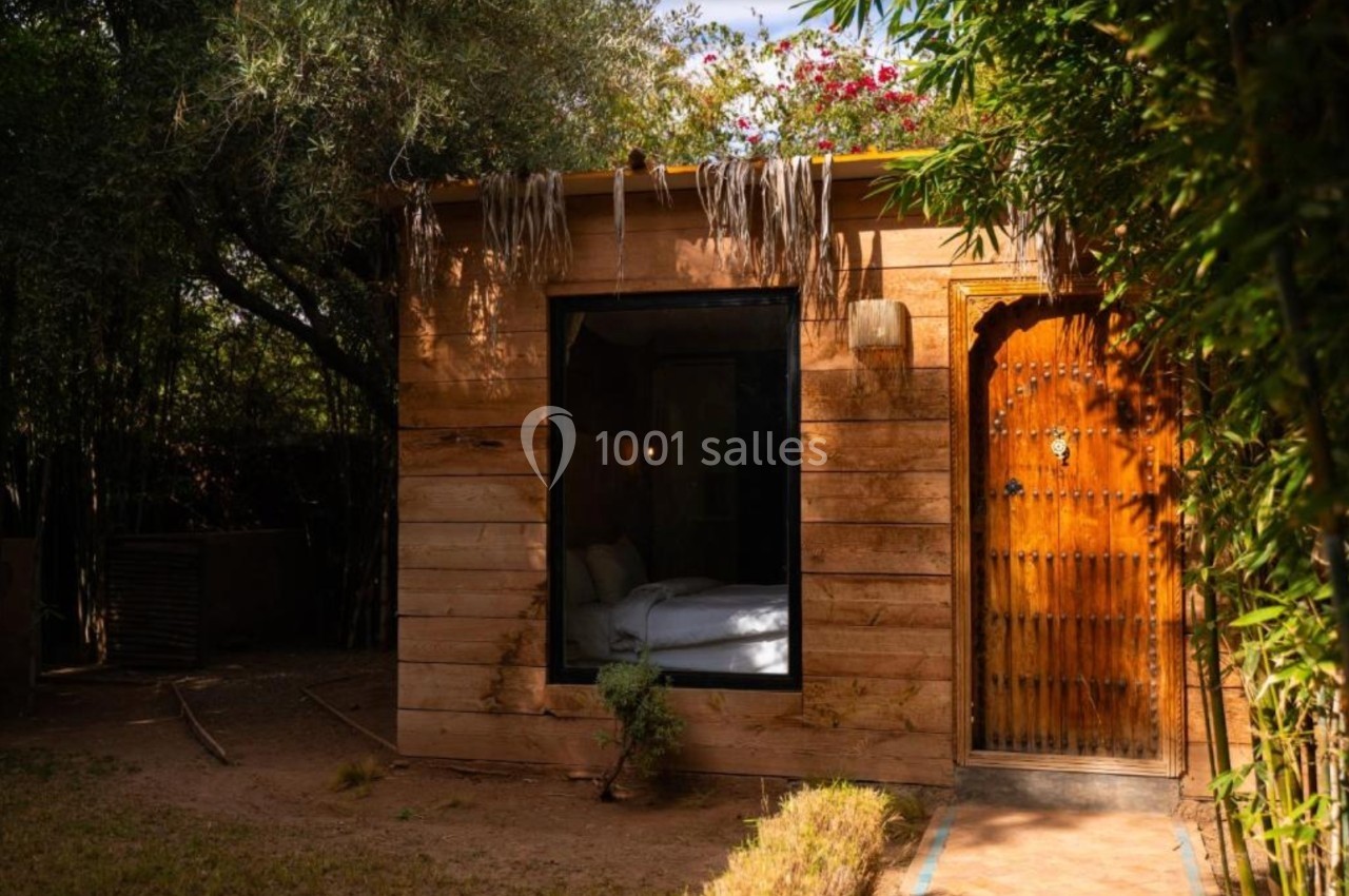 Petite maison en bois avec une porte sculptée, une grande fenêtre et un toit végétalisé, entourée de verdure.