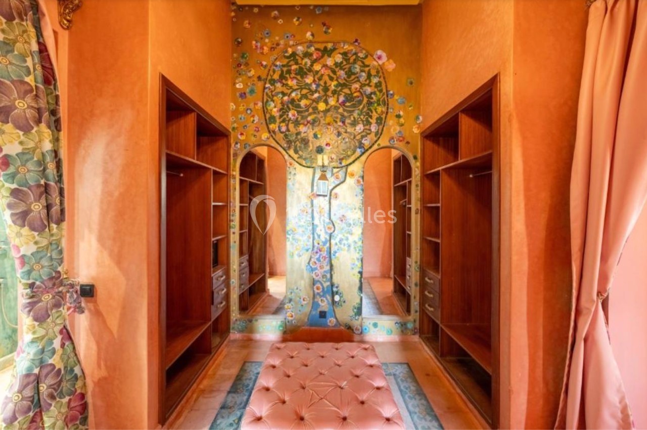 Dressing avec étagères en bois, murs décorés d'une fresque d'arbre fleuri et pouf capitonné rose au centre.