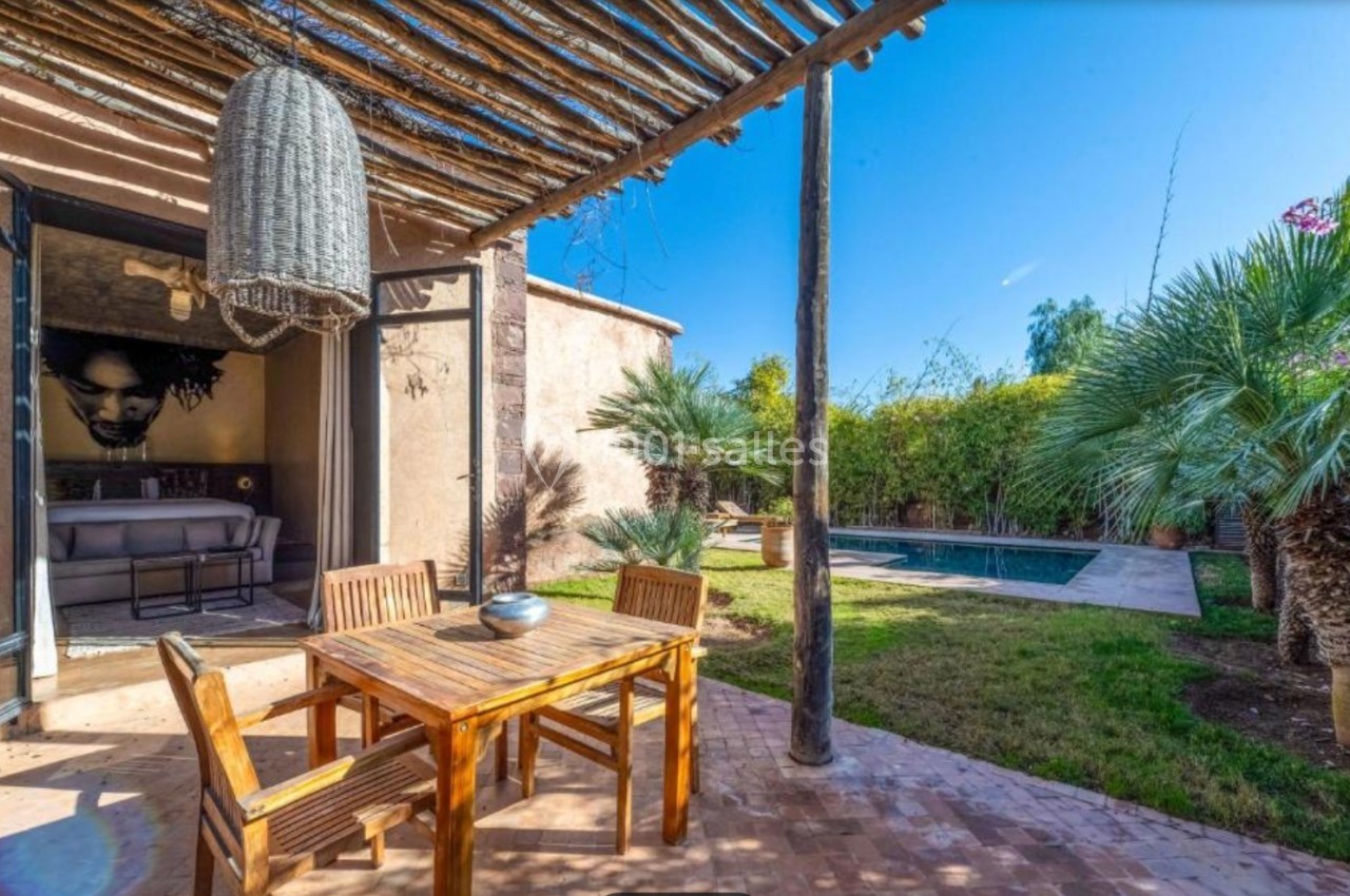 Terrasse avec table en bois, pergola en bambou, vue sur un jardin avec piscine et palmiers sous un ciel bleu.