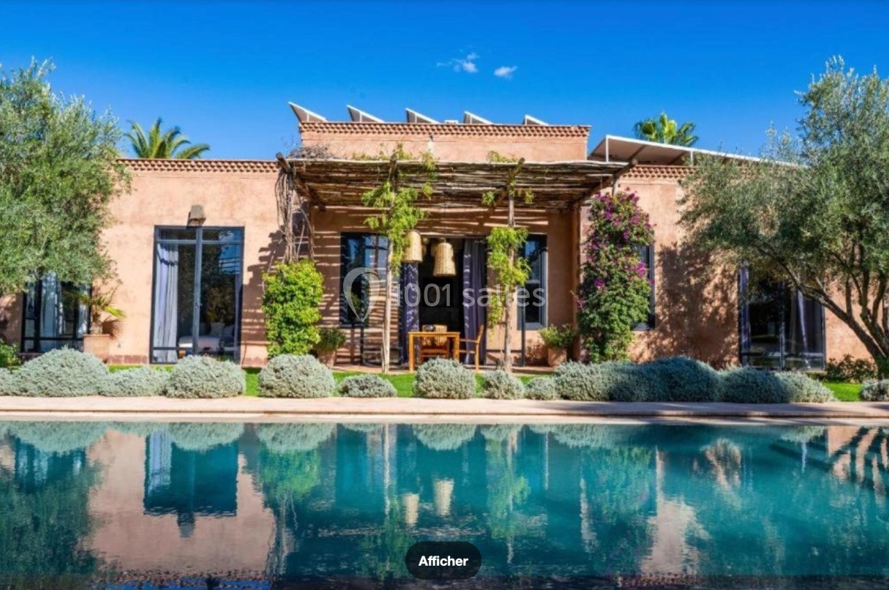 Façade d'une maison en terre cuite avec pergola végétalisée, entourée d'arbres, reflétée dans une piscine calme.