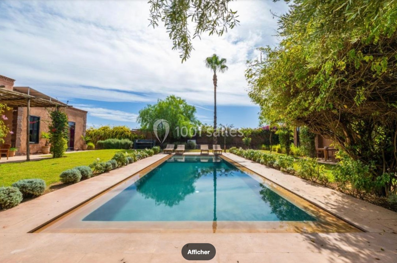 Piscine extérieure entourée de végétation, avec des chaises longues et un ciel partiellement nuageux en arrière-plan.