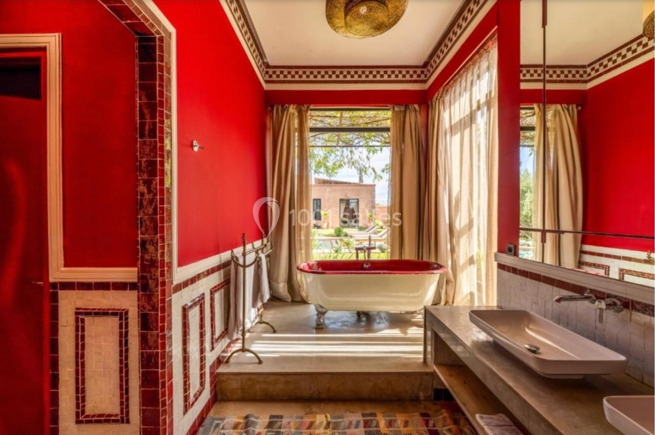 Salle de bain lumineuse avec murs rouges, baignoire sur pieds près d'une fenêtre donnant sur un jardin ensoleillé.