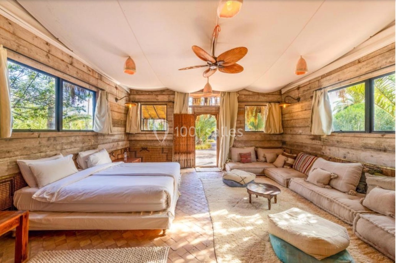 Chambre lumineuse avec murs en bois, lit double, canapés, tapis et grandes fenêtres donnant sur un jardin.