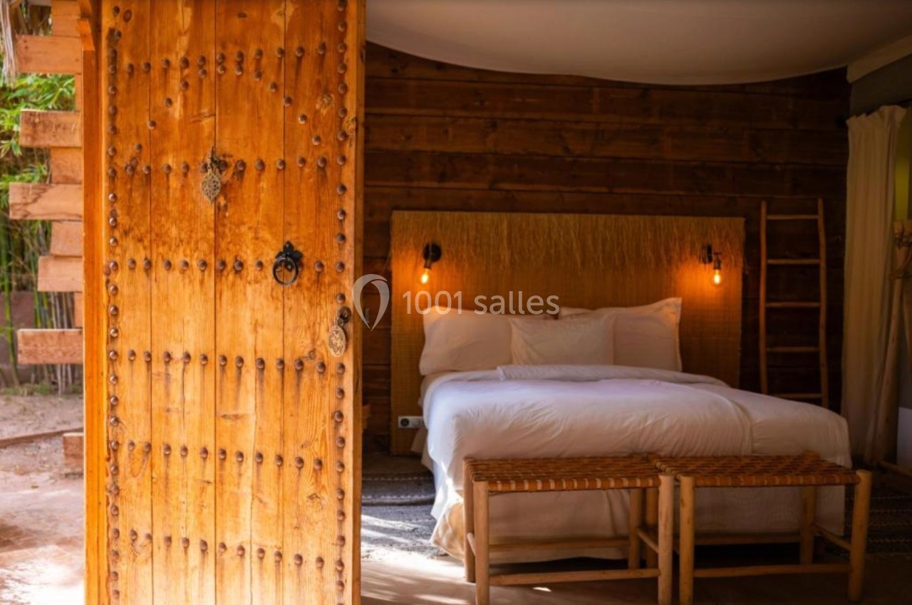 Chambre rustique avec lit double, tête de lit en fibres naturelles, éclairage chaleureux et porte en bois sculpté.