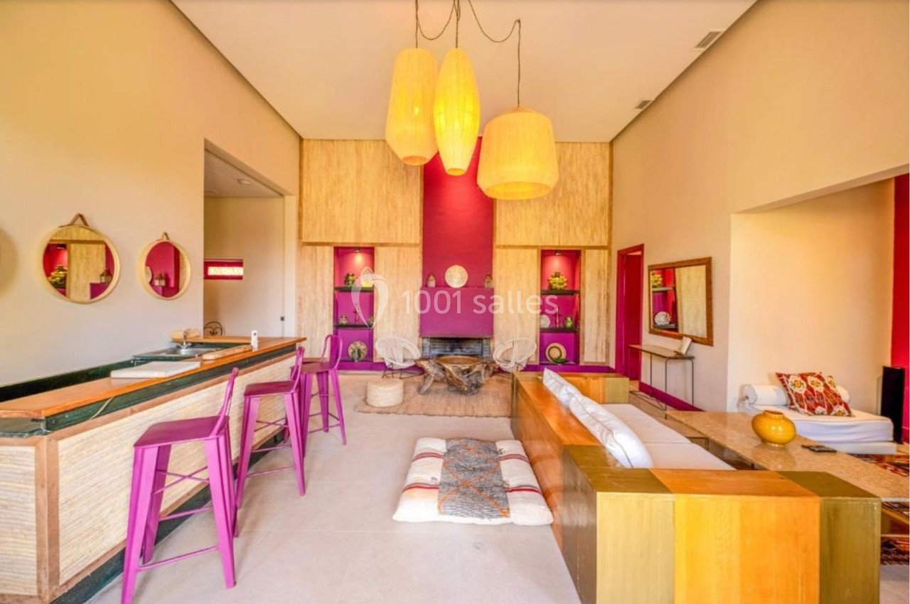 Salon coloré avec murs beige et accents violets, mobilier en bois, bar avec tabourets et suspensions lumineuses.