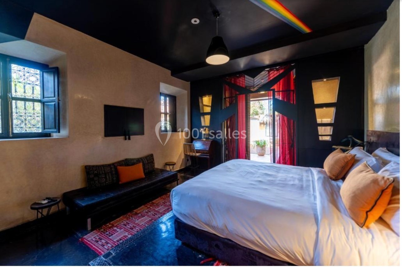 Chambre lumineuse avec lit double, canapé, bureau, télévision, et porte-fenêtre donnant sur une terrasse.