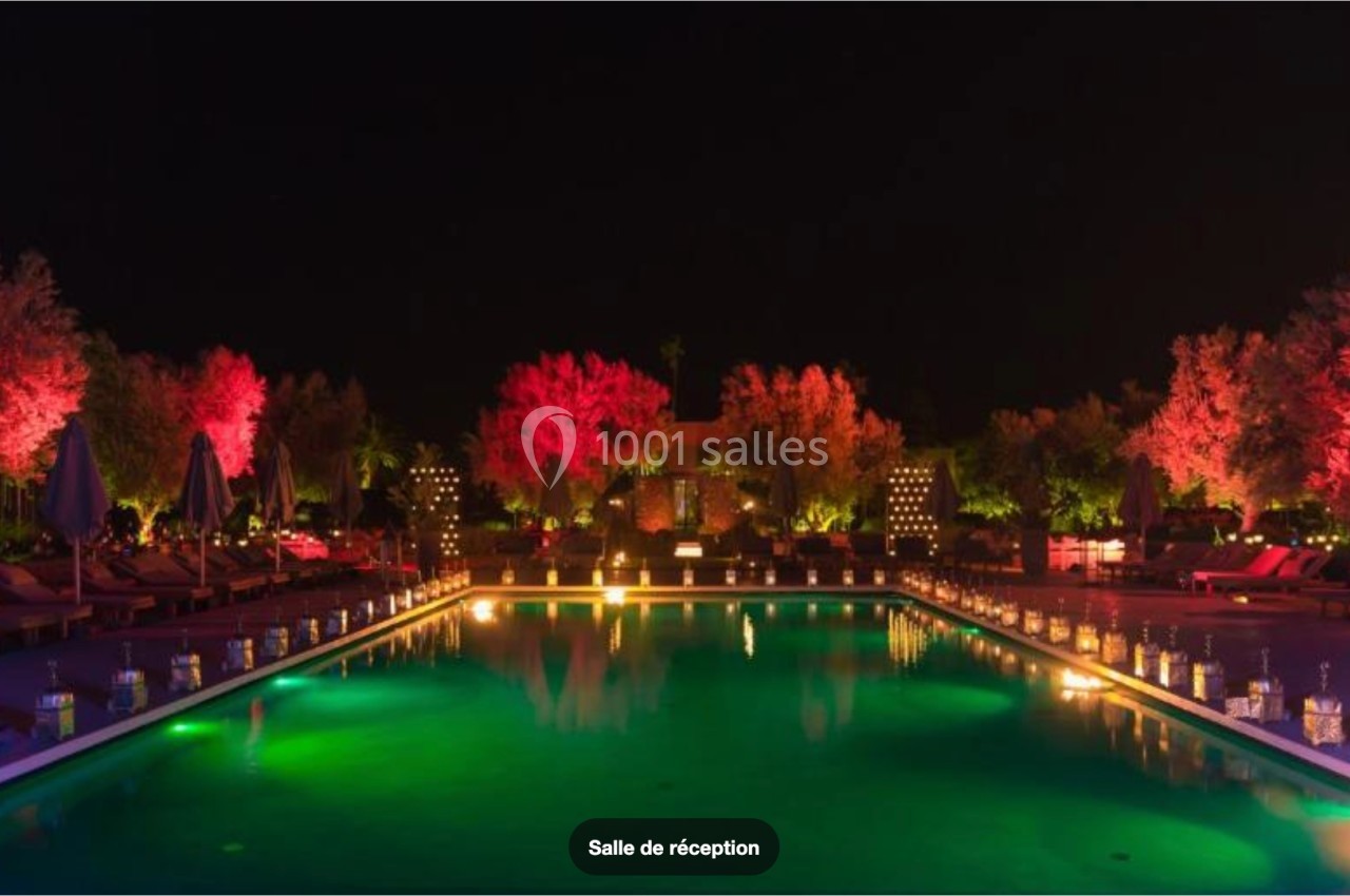Piscine éclairée de nuit entourée de chaises longues, d'arbres illuminés et de lanternes au bord de l'eau.