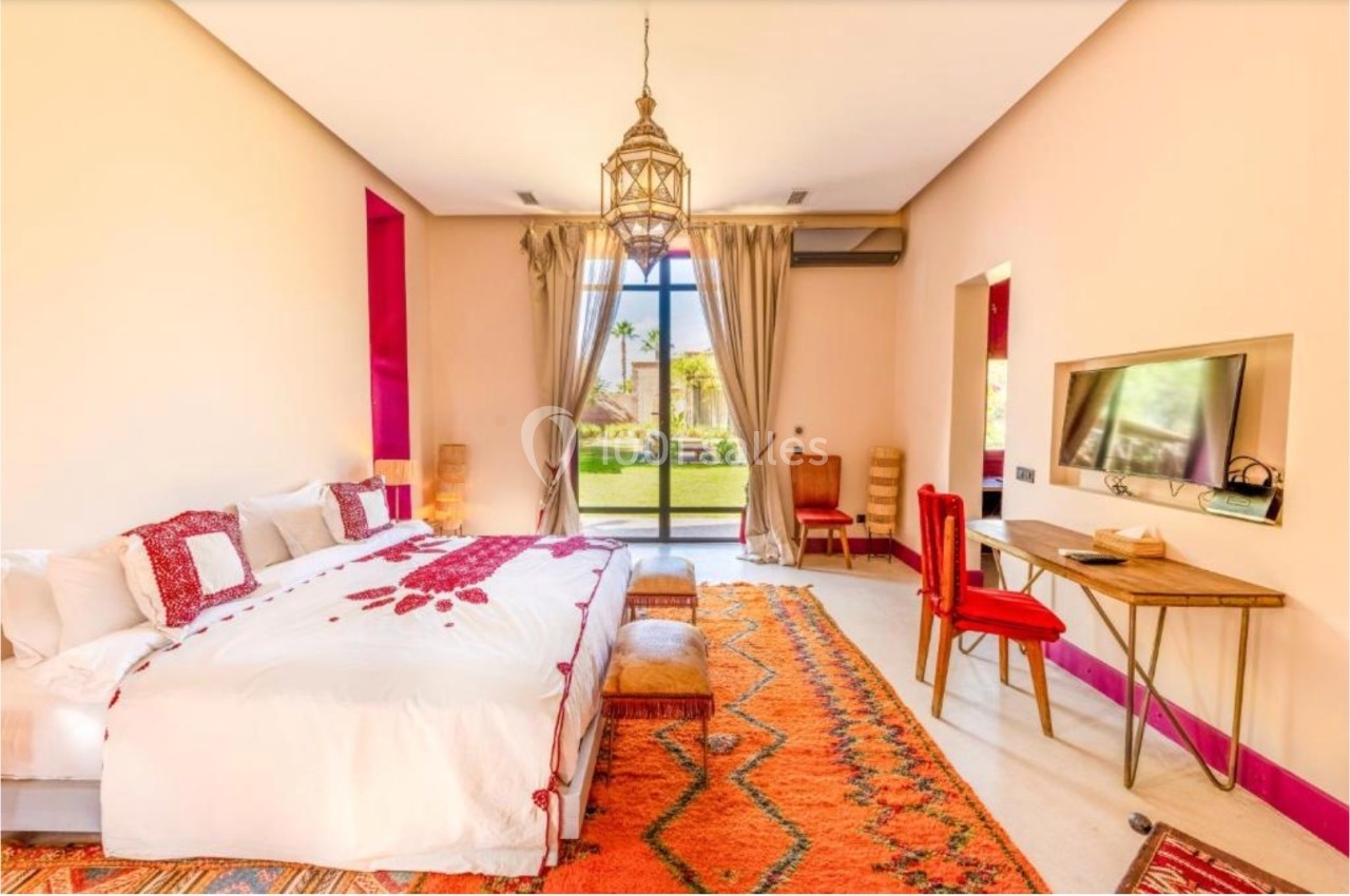 Chambre lumineuse avec lit double, tapis coloré, bureau, télévision et grande baie vitrée donnant sur un jardin.