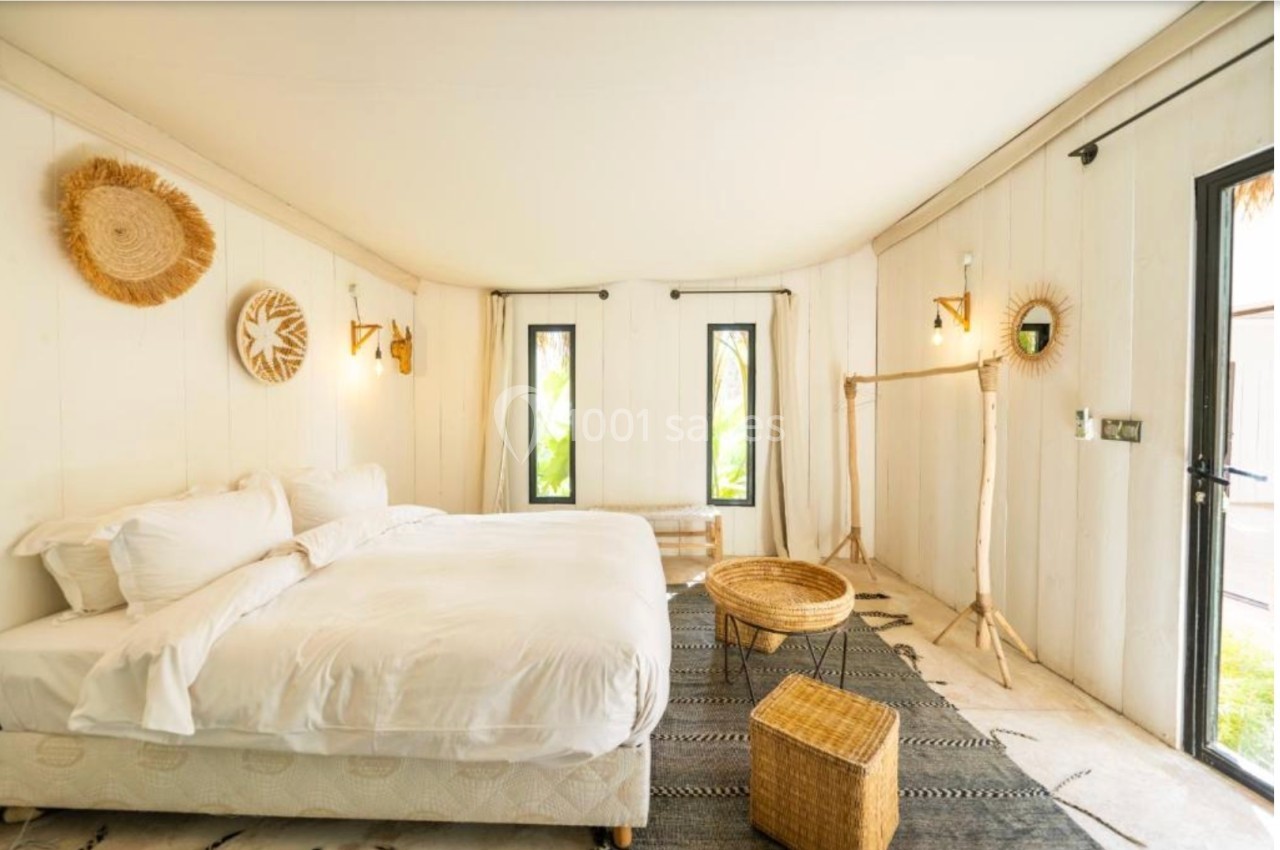 Chambre lumineuse avec lit double, décoration en fibres naturelles, tapis au sol et fenêtres donnant sur un jardin.