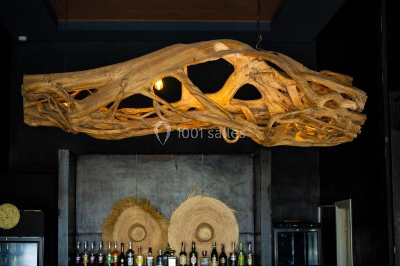 Suspension en bois sculpté avec ampoules intégrées, suspendue au plafond d'un bar sombre avec des bouteilles en arrière-plan.