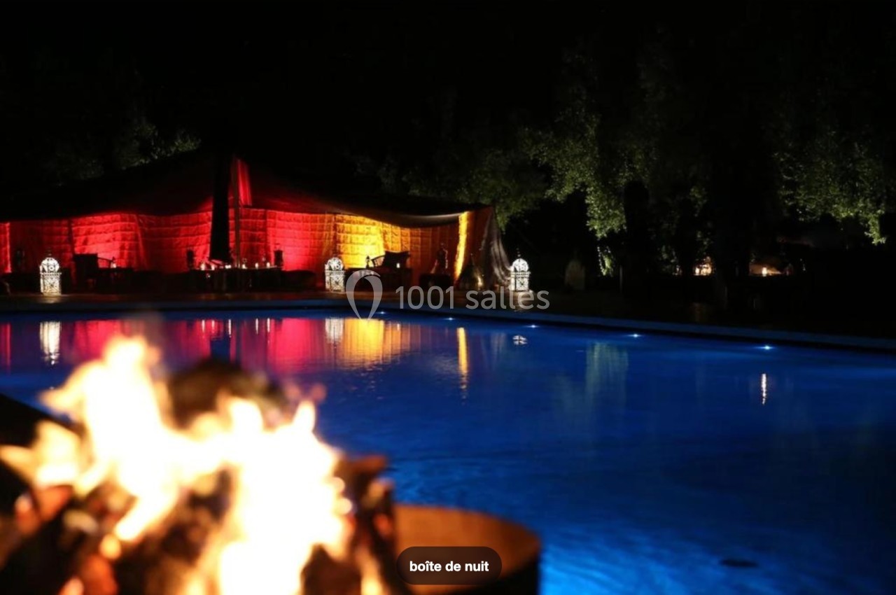 Feu de camp en premier plan avec une piscine éclairée et une tente illuminée en arrière-plan, de nuit.