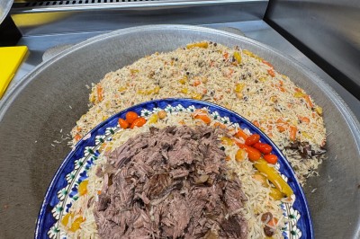 Un homme cuisine de grandes pièces de viande dans une marmite fumante en extérieur, portant un tablier et des gants.