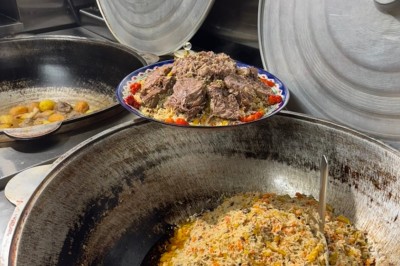 Un homme cuisine de grandes pièces de viande dans une marmite fumante en extérieur, portant un tablier et des gants.