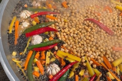 Un homme cuisine de grandes pièces de viande dans une marmite fumante en extérieur, portant un tablier et des gants.